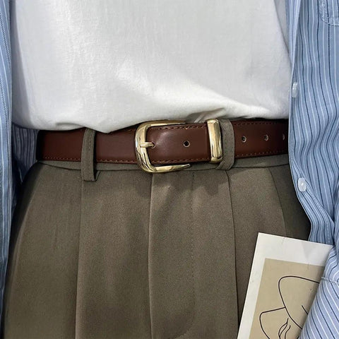 Person wearing a brown belt with a gold metal buckle over gray pants and a white shirt
