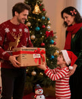 Family exchanging Christmas gift box in front of decorated tree, wearing festive sweaters and Santa hat, smile on child's face.