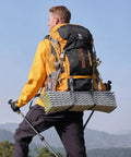 Person hiking with a 65L Internal Frame Hiking Backpack with rain cover, showing nylon material, in outdoor setting, wearing trekking poles.