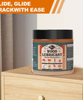 Premium Wood Lubricant Paste Wax That Unsticks Stubborn Drawers And Cabinet Slides By Cutting Friction From Antique Furniture Joints With Zero Residue Comes Ready With Application Brush And Work Gloves on a wooden dresser near curtains.