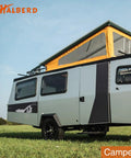 Side view of a modern camper trailer with pop-up roof, featuring black and gray design, parked on grass under a blue sky.
