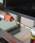 Close-up view of MT2 pen mandrel in wood lathe; 7mm drill bit cutting orange wood.
