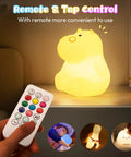 Remote control beside glowing Kids Night Light With Remote, 7 Color Kawaii Cat Nursery Lamp on a nightstand.