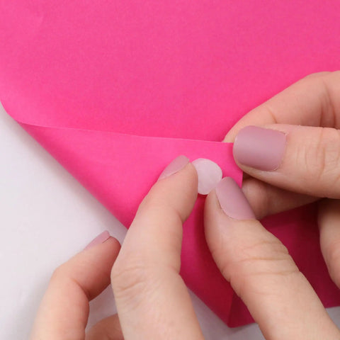 Reusable Mounting Adhesive Putty Dots Pack Of 100 Colorless For Posters, Artwork And Certificates At Home, In Class Or Dorm, Easy To Use, Quick Hold, Removable And Non Messy, Leaves No Residue On Surfaces, white adhesive dot held between fingers on pink paper