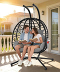 Couple sitting on oversized double hanging egg chair with stand for indoor outdoor use 2 person wicker swing chair with cushions