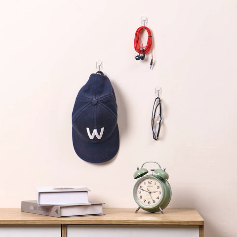 Movelyst clear adhesive wall hooks with strips; front view holds navy cap, red earphones, and glasses.