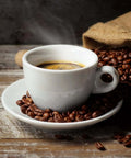 Steaming cup of coffee in white cup on saucer surrounded by roasted coffee beans spilling from burlap sack on wooden table