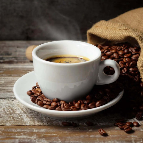 Steaming cup of coffee in white cup on saucer surrounded by roasted coffee beans spilling from burlap sack on wooden table