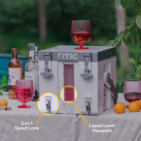 RTIC cooler at outdoor party with glass on top, featuring 3-in-1 spout lock and liquid level viewport, surrounded by bottles and glasses.