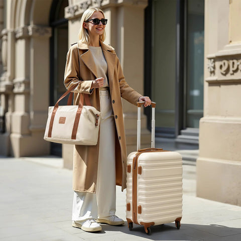 Luggage set: cream hard-shell carry-on suitcase with tan trim and beige duffel bag, carried by a woman in a camel coat.