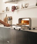 Side view of Ooni 12-inch aluminum pizza peel with black handle placing pizza into a compact countertop oven