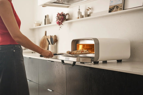 Side view of Ooni 12-inch aluminum pizza peel with black handle placing pizza into a compact countertop oven