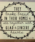Wooden sign with quote from Acts 2:46 surrounded by a vine design, displayed on a wooden background with green plants.