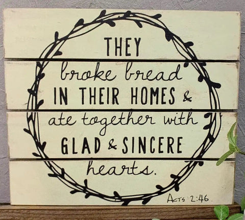 Wooden sign with quote from Acts 2:46 surrounded by a vine design, displayed on a wooden background with green plants.
