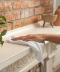 Microfiber cleaning cloth, white, being used to wipe a white mantel against a brick wall.