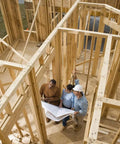 Three people reviewing construction plans inside a wooden house framework under construction.