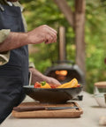 Person cooking vegetables in a pan with a wooden cutting board, utensils, and bowls on an outdoor kitchen counter.