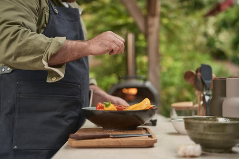 Person cooking vegetables in a pan with a wooden cutting board, utensils, and bowls on an outdoor kitchen counter.