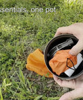 Hands holding Titanium 1100ml Pot And Pan Set For Backpacking with contents inside, mesh bag on grass, outdoor setting