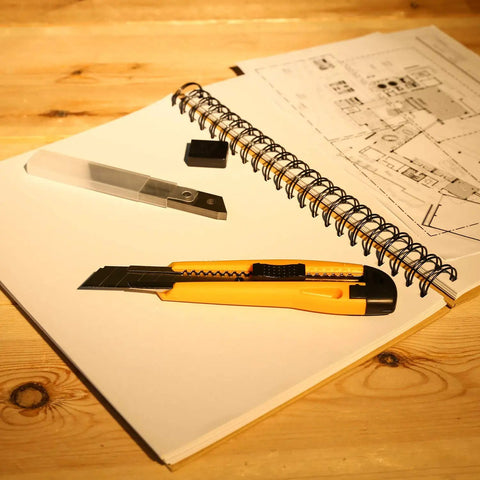 Yellow utility knife and spare blades on notebook with architectural drafts, for 208 Piece General Household Hand Tool Kit With Plastic Storage Case Auto And Home Repair Ready DIY Must Have Set For Hanging Pics, Tightening Screws, Basic Fixes Includes Hammer, Screwdrivers, Pliers, Wrenches.