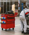 Mechanic using a red tool cart with Magnetic Spray Can Holder Set Six Pack Wall Mount Rack For Spray Cans Paint Cans Bottles And Cleaners Easy Access Tool Organizer For Garage And Home Workshop Black Finish attached on the side in a garage setting.