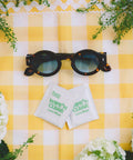 Front view: Flents Wipe N Clear Lens Wipe packets (2) beside black tortoiseshell sunglasses on yellow checkered surface.