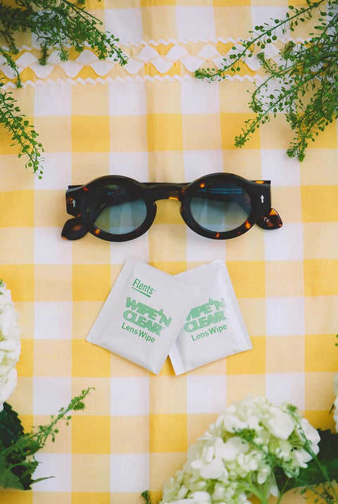 Front view: Flents Wipe N Clear Lens Wipe packets (2) beside black tortoiseshell sunglasses on yellow checkered surface.