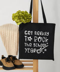 Black tote bag with 'Get ready to rock the school year' silver print, displayed with a sweater, shoes, and green plant for decor.