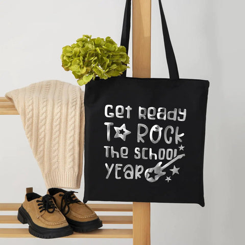 Black tote bag with 'Get ready to rock the school year' silver print, displayed with a sweater, shoes, and green plant for decor.