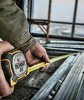 Person using 25 ft tape measure with 1-1/4 inch tape for accurate measurements in carpentry project, visible framing structure
