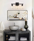 Three Light Black Vanity Lights Over Mirror, On Trend Modern Farmhouse Industrial Bathroom Fixtures With Metal Base And Clear Glass Shades, Wall Sconce Lighting For Bathrooms, Bulbs Not Included above a mirror and vanity setup.