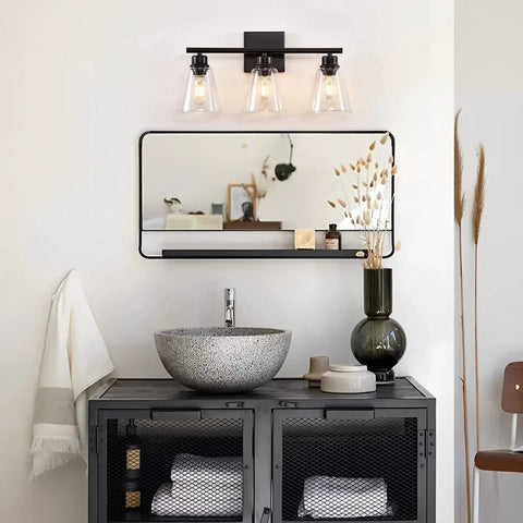 Three Light Black Vanity Lights Over Mirror, On Trend Modern Farmhouse Industrial Bathroom Fixtures With Metal Base And Clear Glass Shades, Wall Sconce Lighting For Bathrooms, Bulbs Not Included above a mirror and vanity setup.