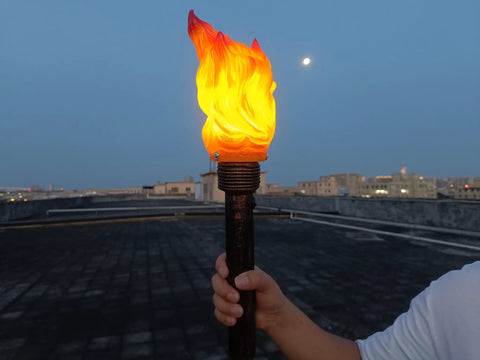 Hand holding a Realistic Flameless LED Torch Light Prop Lamp For Parties, Holidays, Patio Decor, Backyard Fun And Outdoor Sports Activities, Easy To Setup Handheld Style, Battery Powered, Eco Friendly, No Fuel Or Smoke Or Wiring against a cityscape at dusk.