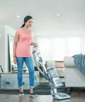 Person vacuuming in a stylish living room with a modern, silver vacuum cleaner on a wooden floor with a cozy sofa.