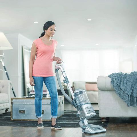 Person vacuuming in a stylish living room with a modern, silver vacuum cleaner on a wooden floor with a cozy sofa.