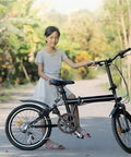 Young girl standing with a black folding bicycle on a sunny road, surrounded by lush greenery.