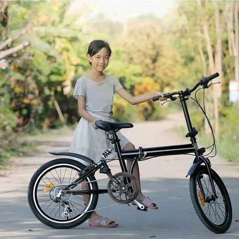 Young girl standing with a black folding bicycle on a sunny road, surrounded by lush greenery.