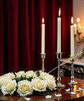 Pretty Slick Gold Metal Candle Snuffer With Extra Long Handle Steady Grip Wick Extinguisher Tool No Burn Drama Heavy Steel Finish Cozy Hearth Decor Perfect Gift For Housewarmers next to lit candles and white roses on a table.