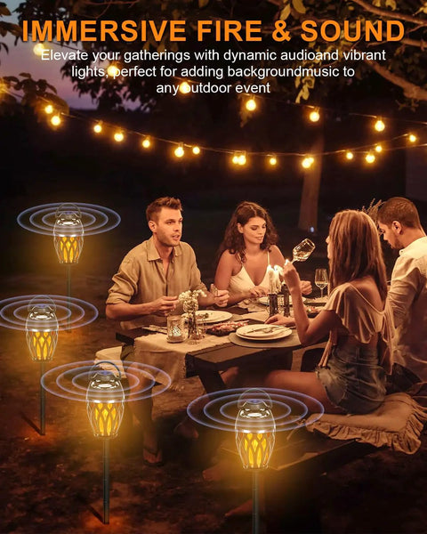 People dining outdoors with Twin Pack Outdoor Bluetooth Flame Speaker With LED Torch Dope Ambience Light, creating a cozy atmosphere.