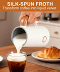 Pouring foam from Powerful 4 In 1 Electric Milk Frother And Warmer With Dual Whisks into coffee with croissant nearby on table.