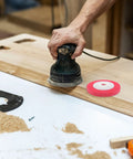 Hand using an electric sander on wood with a red Multipurpose Nylon Grinding Wheel for Metal Wood and Plastic nearby.