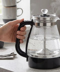 1 Liter Glass Electric Kettle With Keep Warm Function, Rapid Boil Tech, Auto Shut Off Safety, Bpa-Free Borosilicate Build, And An Easy Pour Spout For Your Daily Must Have Morning Routine, held in hand with steam rising.