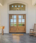 Wooden double front door with glass panels, surrounded by a white wall, with a rocking chair and flower vase on the porch.