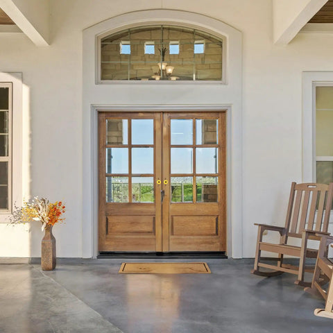 Wooden double front door with glass panels, surrounded by a white wall, with a rocking chair and flower vase on the porch.