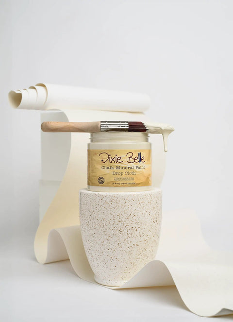 Drop Cloth Chalk Mineral Paint For Furniture In Matte Linen White 16 Oz with paintbrush on a textured surface