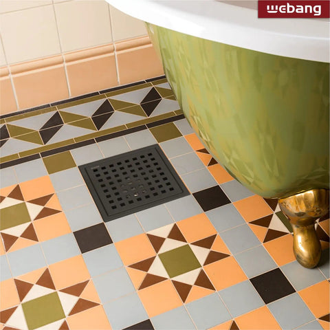 Floor view showing a 6 Inch Square Shower Floor Drain With Flange And Removable Grate Matte Black Stainless Steel SUS 304 Watermark And CUPC Certified Easy Install Clog Resistant Drainage For Fast Water Removal In Standard No Hub Base next to a green bathtub on patterned tiles.