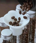 Single Dose Coffee Bean Storage With One Way Exhaust Valve And Metal Stand 14 Piece Glass Jars, pouring beans into clear jar with funnel.