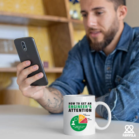 Man holding phone with Funny Engineer Attention Mug For Engineers Gift Coffee Cup 11 Oz White Ceramic Novelty Drinkware in foreground.