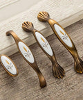 Vintage Bronze Cabinet Handles And Drawer Knobs Set For Kitchen And Furniture No Fuss Install Durable Alloy Hardware With Rust Proof Finish Retro Look For Cabinets Drawers Dress Up Any Room, featuring oval white ceramic inlays with floral design, displayed on wooden surface.