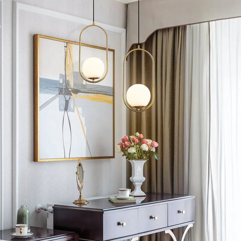 Modern One Light Globe Pendant With White Milk Glass Shade In Brushed Brass Finish For Kitchen Island Farmhouse Living Spaces Mid Century Style Ceiling Lighting Fixture in interior view next to artwork and flowers.